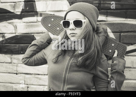 Teenage girl in sunglasses détient plus de skateboard urban mur de briques, l'arrière-plan photo monochrome Banque D'Images