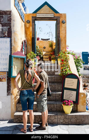 Santorin, Théra. Oia. Sigalas, extérieur, restaurant grec avec des marches menant à l'entrée principale Young tourist couple, homme et femme, la lecture meue. Banque D'Images