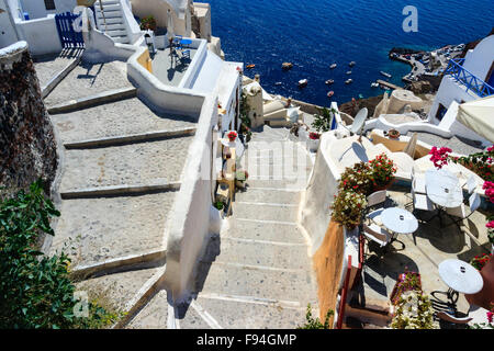 Santorin, Théra. Vue de dessus des marches en pierre se tordant vers le bas pour le bleu de la mer et le port de Oia avec terrasse avec table et chaises blanc mis en place. Banque D'Images