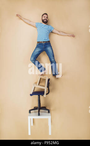 Homme debout dangereusement sur une pile de chaises. Studio shot sur un fond beige. Banque D'Images