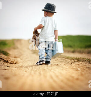 Vue arrière du petit garçon marche sur le trottoir en champ avec suitacase et ours Banque D'Images
