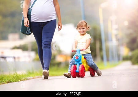 Jeune mère enceinte avec sa petite fille sur petite moto de passer du temps ensemble dans le parc Banque D'Images