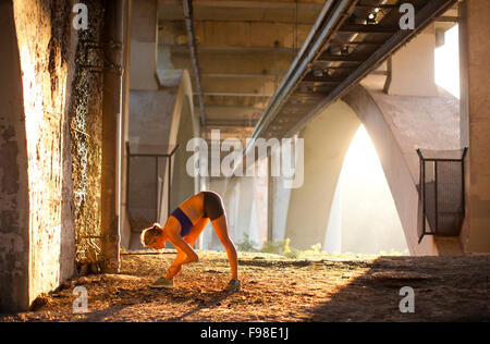 Une femme s'étend avant une course à la lumière du soleil du matin en milieu urbain sous un pont. Banque D'Images