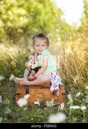 Piscine d'été portrait of cute little girl sitting on meadow avec vieille valise avec des jouets Banque D'Images