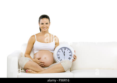 Studio portrait of pregnant woman holding big wall clock assis sur canapé isolé sur fond blanc Banque D'Images