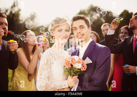 Les jeunes mariés profiter de moment romantique ensemble à réception de mariage à l'extérieur, les invités du mariage en arrière-plan blowing bubbles Banque D'Images