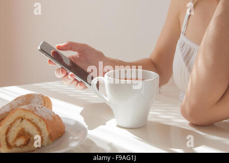 Matin de la belle jeune femme. close-up d'une main de la jeune fille avec un téléphone et une tasse de thé revigorante tôt le matin dans la chambre à coucher Banque D'Images