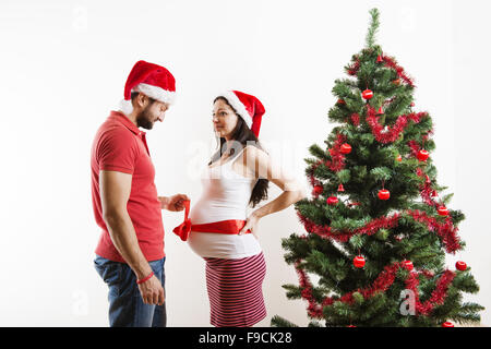 Jeune couple est debout près d'arbre de Noël. Femme est enceinte et attachées avec un ruban rouge. Banque D'Images