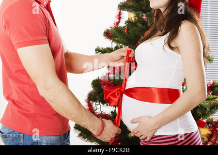 Jeune couple est debout près d'arbre de Noël. Femme est enceinte et attachées avec un ruban rouge. Banque D'Images