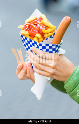 Les mains tenant le cône de papier avec une saucisse chaude et de frites avec du ketchup Banque D'Images