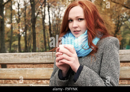 Redhead girl avec tasse de thé s'asseoir sur un banc de parc de la ville, saison d'automne Banque D'Images