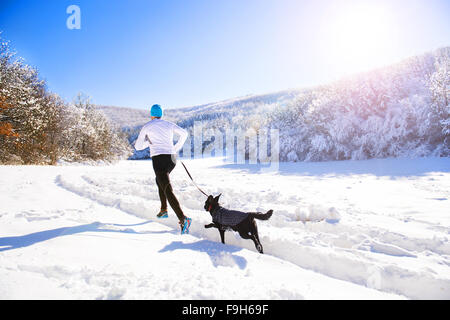 Jeune sportif jogging avec chien à l'extérieur dans le parc d'hiver ensoleillé Banque D'Images