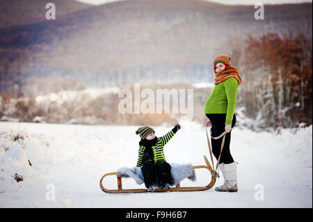 Maman heureuse avec son fils à cheval sur un traîneau s'amusant à l'extérieur dans la neige. Banque D'Images