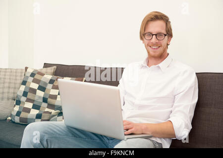 Jeune homme aux cheveux rouges avec des lunettes assis sur table avec ordinateur portable et sourit à huis clos Banque D'Images