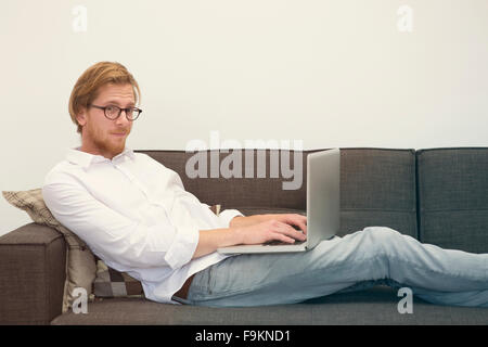 Jeune homme aux cheveux rouges avec des lunettes sitting on couch with laptop Banque D'Images
