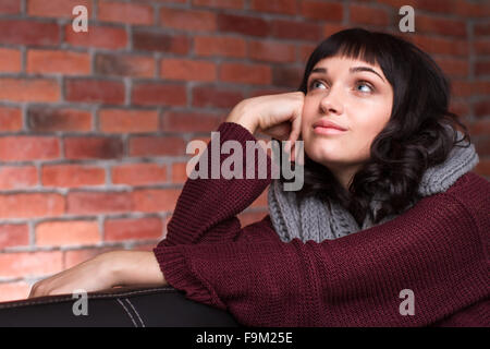 Jolie jeune femme en vêtements tricotés assis et rêver sur fond brique Banque D'Images