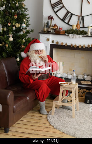 Santa Claus boire du lait et de la lecture livre près de cheminée à la maison Banque D'Images