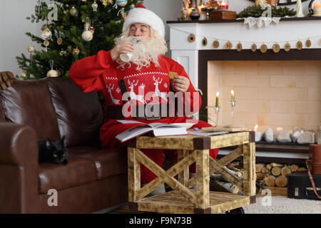 Santa Claus boire du lait et de la lecture livre près de cheminée à la maison Banque D'Images