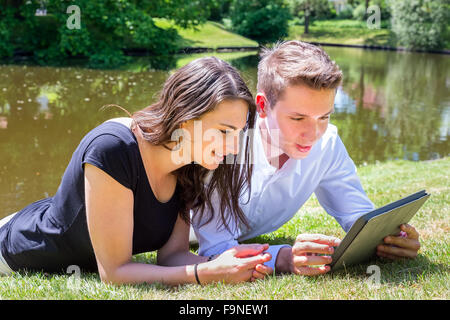 Jeune couple lying on grass à la lecture de l'eau l'Ipad Banque D'Images