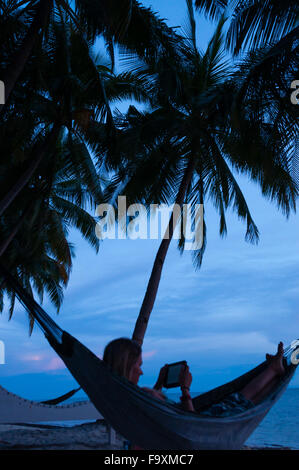 Femme lisant sous les palmiers portant dans un hamac sur la plage pendant le coucher du soleil Banque D'Images