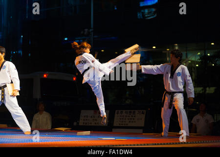 Une ceinture noire de Taekwondo une adolescente sautant en l'air des coups de pied et de briser une planche de bois tenu par des collègues à un parking extérieur su Banque D'Images