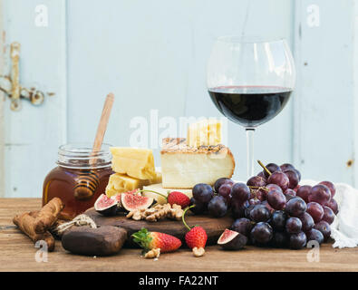 Verre de vin rouge, plateau de fromages, raisins,fig, fraises, miel et bâtonnets de pain sur table en bois rustique, fond bleu, séle Banque D'Images