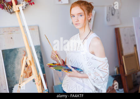 Thoughtful young female painter en utilisant les peintures à l'huile pour la peinture sur toile en atelier d'art Banque D'Images