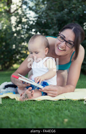 Mère et bébé garçon tenant un livre d'images et de rire en pelouse, Munich, Bavière, Allemagne Banque D'Images