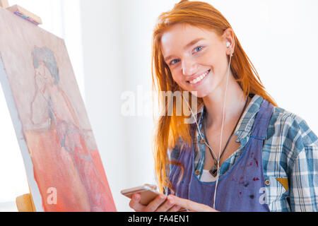 Cheerful attractive rousse jeune femme peintre debout près de photo et écouter de la musique depuis un téléphone portable Banque D'Images