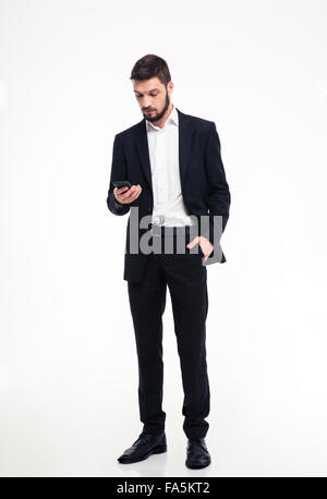 Toute la longueur du jeune homme barbu confiance attrayant en costume noir à l'aide de téléphone sur fond blanc Banque D'Images