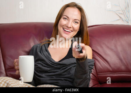 Cheerful attractive young woman sitting in living room with tv remote control et boire le thé sur canapé Banque D'Images