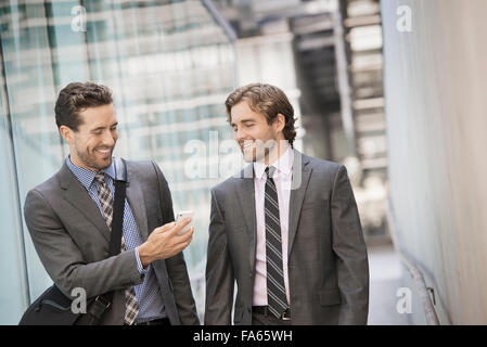 Deux hommes en costumes d'affaires à l'extérieur d'un grand bâtiment, l'un tenant un téléphone intelligent. Banque D'Images