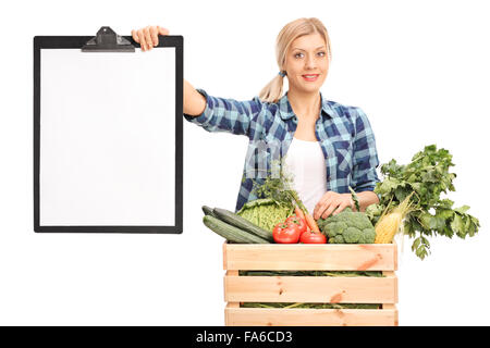 Jeune femme tenant un presse-papiers et debout derrière une caisse pleine de légumes frais isolé sur fond blanc Banque D'Images