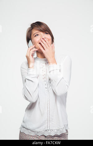 Jolie jeune femme en blouse blanche couvert la bouche avec la main et parler sur smartphone Blackberry sur fond blanc Banque D'Images