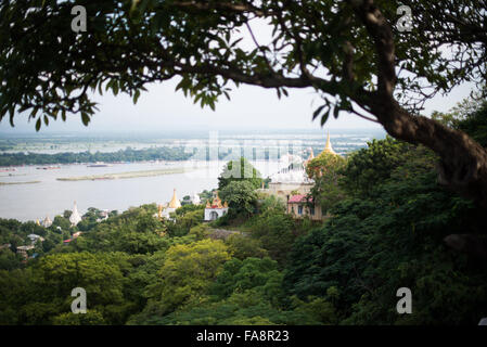 Sagaing Hill Stupas Ayeyarwady River Through Trees Myanmar | SAGAING, Myanmar — Des stupas dorés et un complexe monastique à flèche blanche sur la crête boisée de Sagaing sont encadrés par des branches d'arbres surplombantes, avec le large fleuve Ayeyarwady s'étendant au-delà, vus depuis la terrasse de la pagode Soon U Ponnya Shin. Chaque capitale royale visible depuis ce sommet au fil des siècles — Inwa, Amarapura et Mandalay — a parrainé la pagode à tour de rôle, liant le pouvoir temporel à la prétendue prééminence religieuse de la crête. Banque D'Images