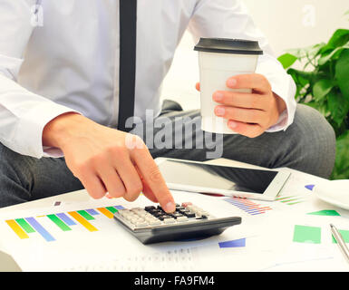 Libre d'un jeune homme de race blanche à l'aide d'une calculatrice sur une table pleine de tableaux et une tasse de café dans sa main Banque D'Images