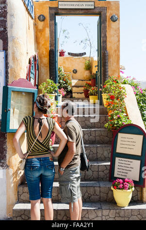 Santorin, Théra. Oia. Sigalas, extérieur, restaurant grec avec des marches menant à l'entrée principale Young tourist couple, homme et femme, la lecture meue. Banque D'Images