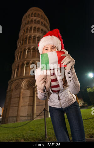 L'emblématique de l'architecture italienne ajoute du style à la célébration de Noël. Happy young woman in Santa hat se cacher derrière un drapeau italien à l'avant de la Tour Penchée de Pise, Italie dans la soirée Banque D'Images