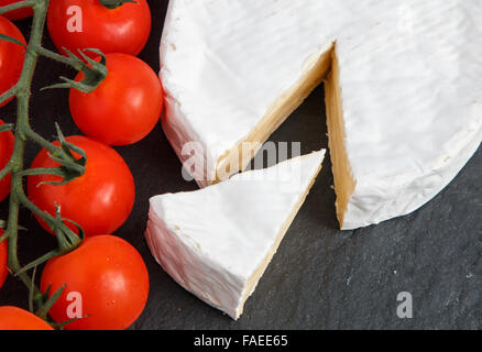 Tranche de Brie coupé d'un fromage entier à côté de tomates cerises rouges sur une base d'ardoise gris Banque D'Images