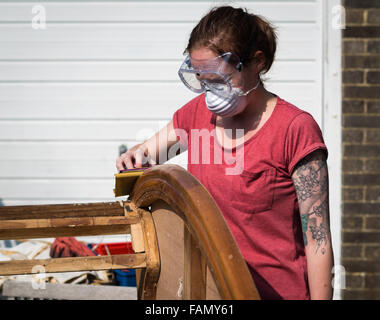 Jeune femme tatouée ponçage à la main un morceau de meubles anciens en bois à l'extérieur portant l'équipement de protection. Banque D'Images