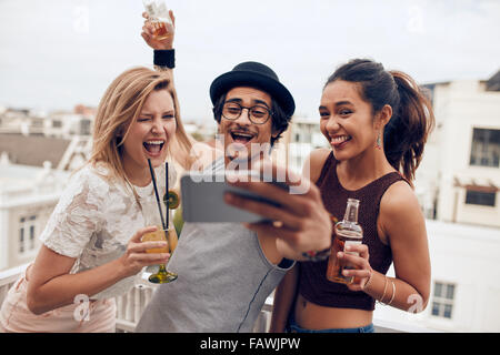 Petit groupe d'amis en tenant sur un selfies téléphone mobile. Jeune homme et femmes avec boissons faire drôle de visage tout en prenant un po Banque D'Images