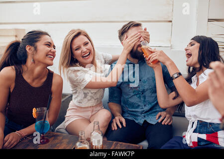 Jeunes amis assis jouissant ensemble partie. Femme fermeture yeux d'un homme avec un autre donner à boire. Les jeunes s'amusant à Banque D'Images