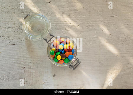 Au chocolat enrobés de sucre coloré dans un bocal en verre sur la table en bois blanc avec des taches sur la table. Banque D'Images