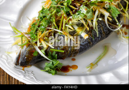 Près d'un poisson entier à la vapeur fraîchement couverts avec des herbes, des oignons et de la sauce sur plaque blanche. Banque D'Images