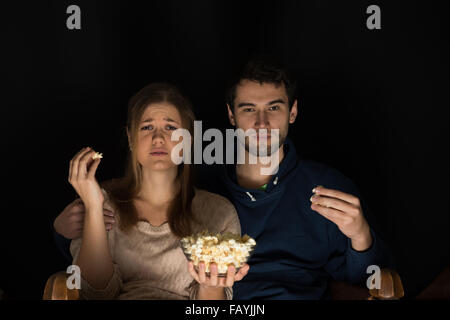 Jeune couple, homme et femme, assise dans la pièce sombre à l'avant de regarder la télévision et film eating popcorn, montrant les émotions Banque D'Images