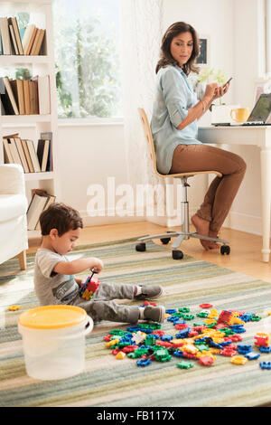 Boy (2-3) Playing with toy blocks alors que mère working on laptop Banque D'Images