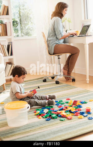 Boy (2-3) Playing with toy blocks alors que mère working on laptop Banque D'Images