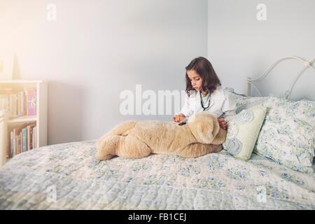 Girl dressed up in lab coat assis au lit en utilisant le stéthoscope soft toy Banque D'Images