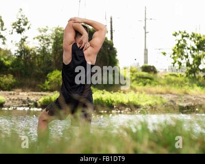 Jeune homme exerçant au bord de la rivière, s'étirant les bras, vue arrière Banque D'Images