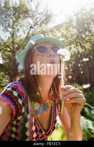 Young woman wearing sunhat et lunettes blowing dandelion seeds Banque D'Images